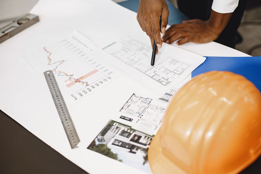 Mãos revisando desenhos técnicos sobre a mesa, ao lado de um capacete de segurança